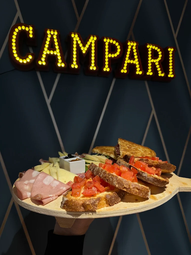 Tagliere di bruschette e salumi servito davanti all'insegna luminosa Campari